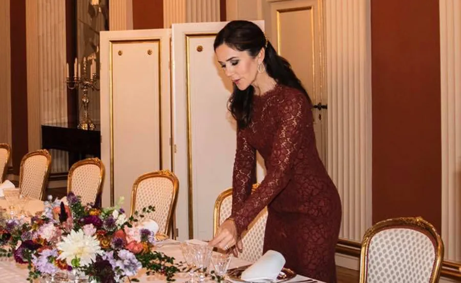 A woman arranges a formal dining table with floral centerpiece and chairs in an elegant room.