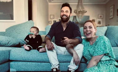 Family sitting on a sofa; a baby with a pacifier, a man with a remote, and a laughing woman in a floral dress.