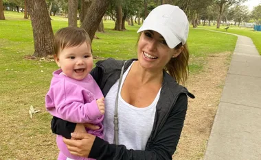 Woman in a white cap holding a smiling baby in a pink outfit at a park.