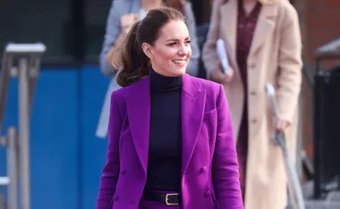 A woman in a vibrant purple pantsuit smiles while standing outside, with people in the background.