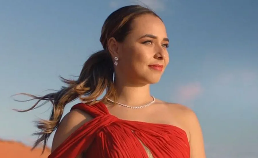 A woman in a red dress with a ponytail stands against a clear blue sky, wearing earrings and a necklace.