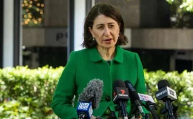 A woman in a green coat speaks at a press conference, surrounded by microphones from various news outlets.