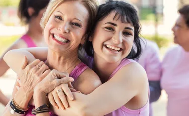 Two women in pink tops smiling and embracing each other outdoors, with others blurred in the background.