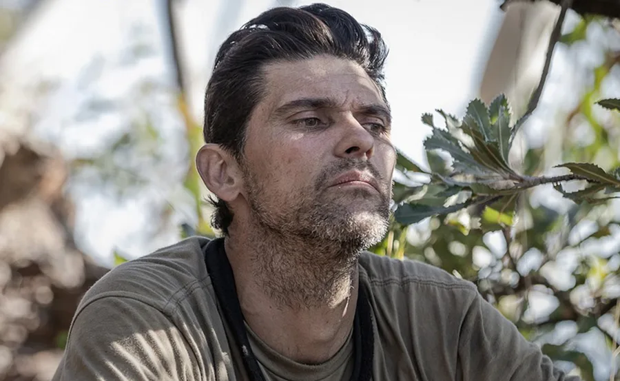 A contemplative man, outdoors, surrounded by foliage, looking into the distance.