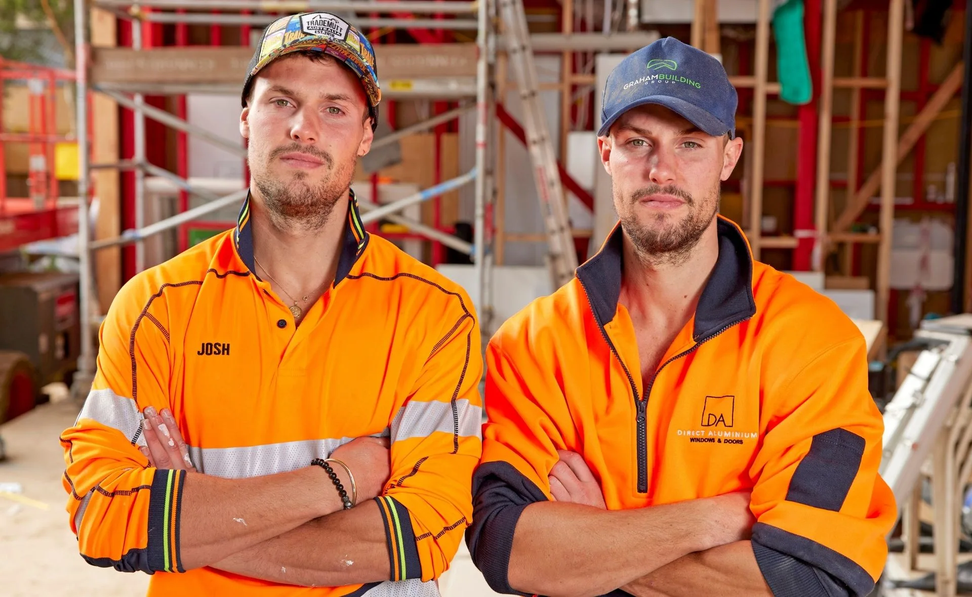 Two men in bright orange workwear with arms crossed stand at a construction site, scaffolding in the background.