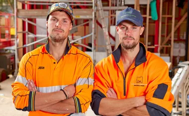Two men in bright orange workwear with arms crossed stand at a construction site, scaffolding in the background.