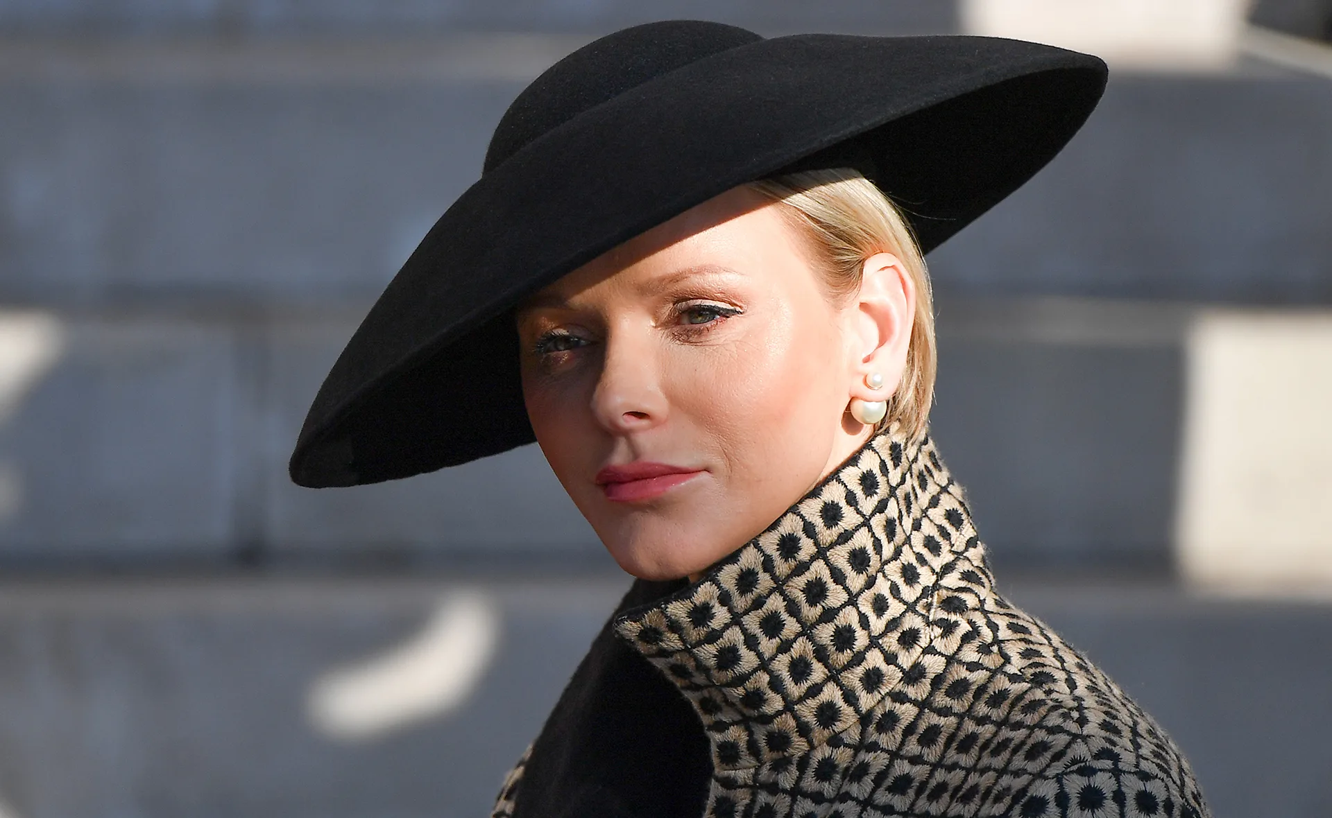 Princess Charlene wears a stylish black hat and patterned coat, looking poised and elegant outdoors.