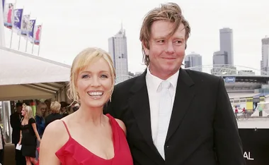 A woman in a red dress and a man in a suit smile together outdoors, with city skyline in the background.