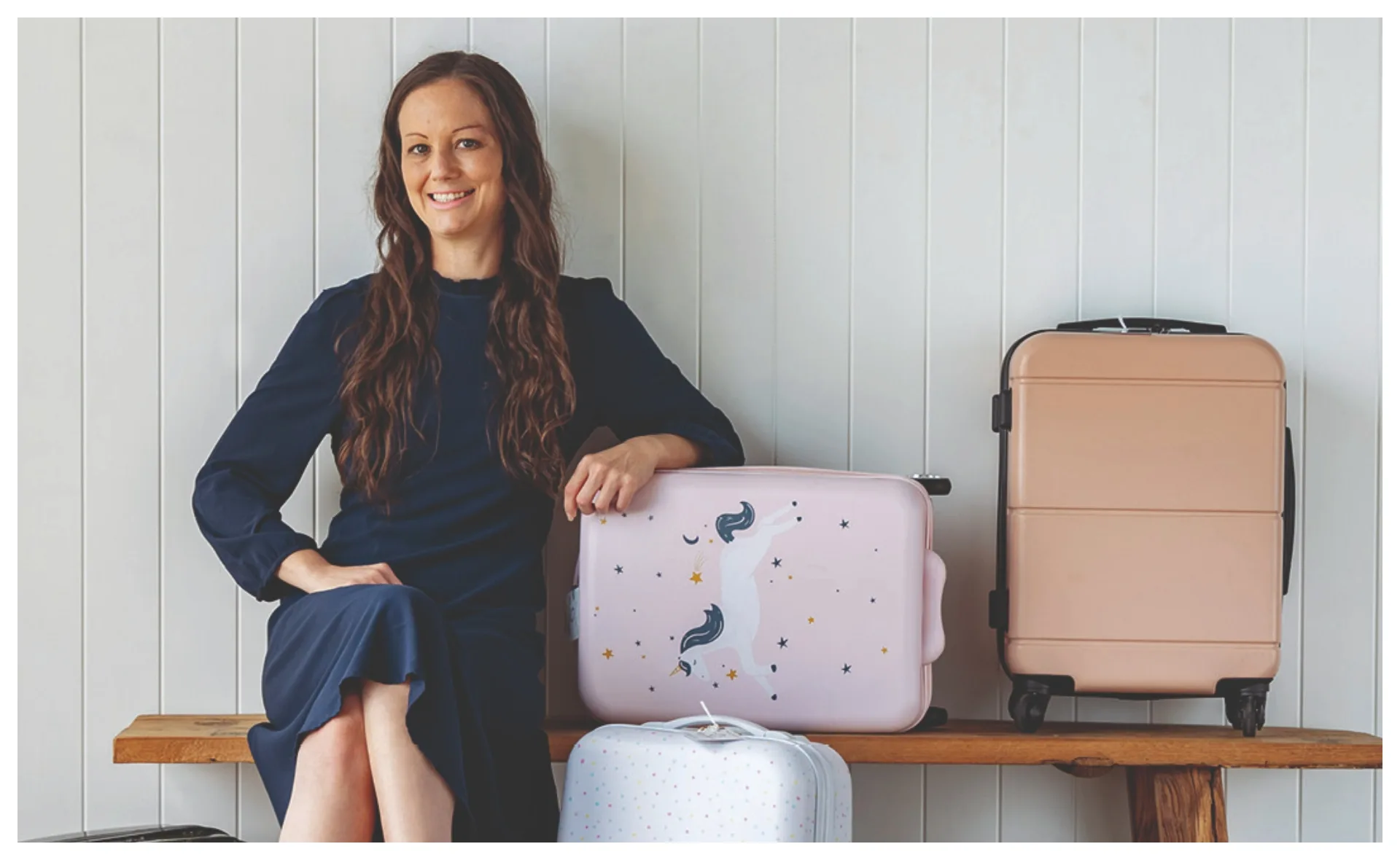 A woman in a navy dress sits on a bench next to three suitcases, one pink with a unicorn design.
