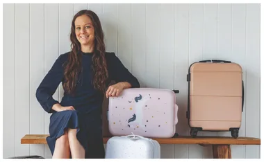 A woman in a navy dress sits on a bench next to three suitcases, one pink with a unicorn design.
