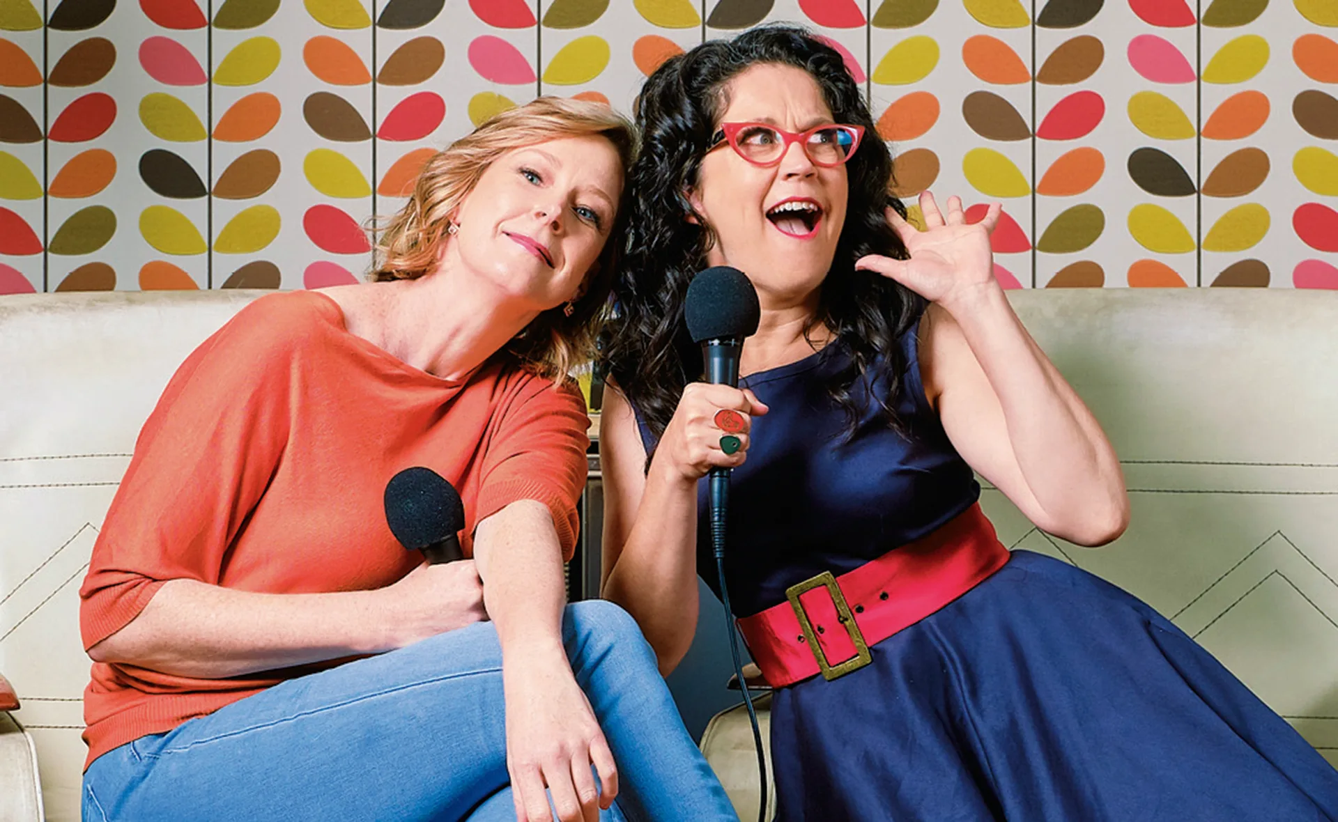 Two women sitting on a sofa, holding microphones, with a colorful leaf-patterned wallpaper background.