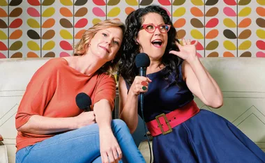 Two women sitting on a sofa, holding microphones, with a colorful leaf-patterned wallpaper background.