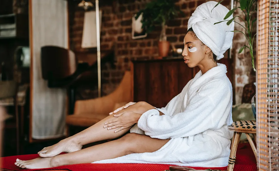 Person in a bathrobe with eye patches and a towel on head, sitting on a red mat in a cozy room.