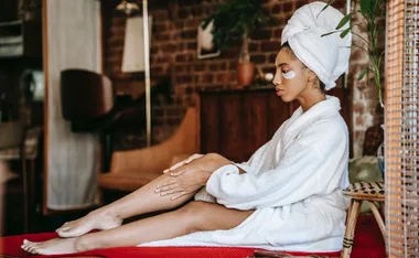 Person in a bathrobe with eye patches and a towel on head, sitting on a red mat in a cozy room.