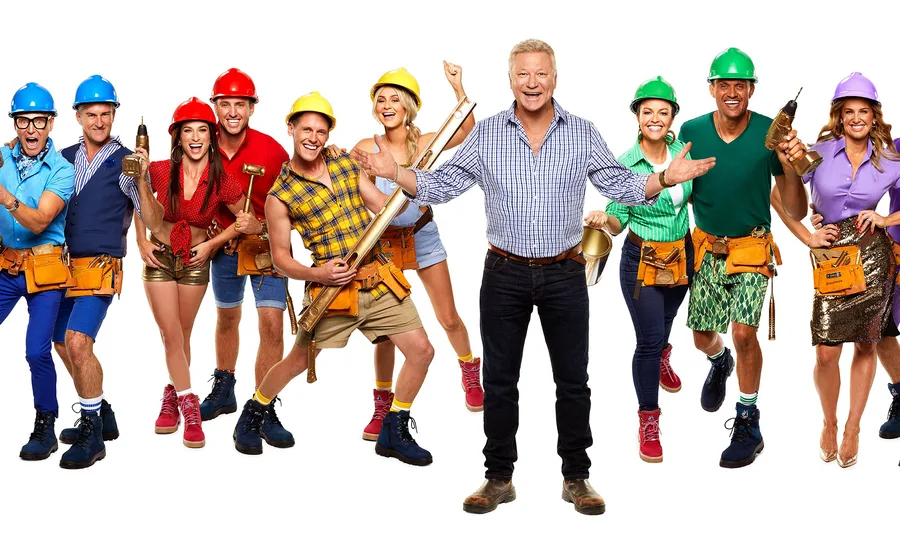 Group of contestants and host from a construction-themed TV show, wearing hard hats and tool belts, smiling.