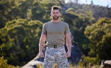 A man in a khaki t-shirt and camo shorts stands confidently outdoors, with trees in the background.