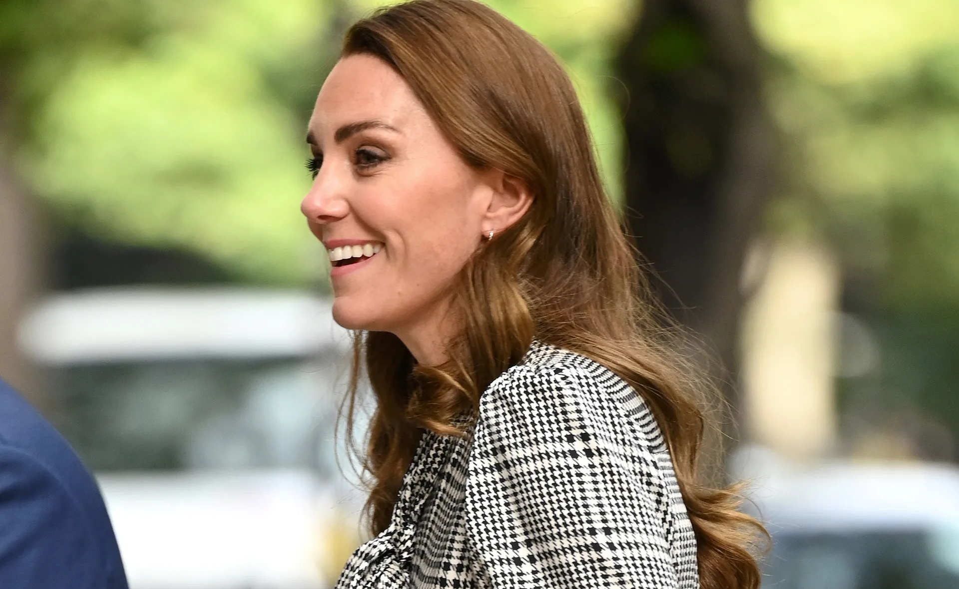 A woman with long brown hair smiles, wearing a black and white houndstooth dress, outdoors.