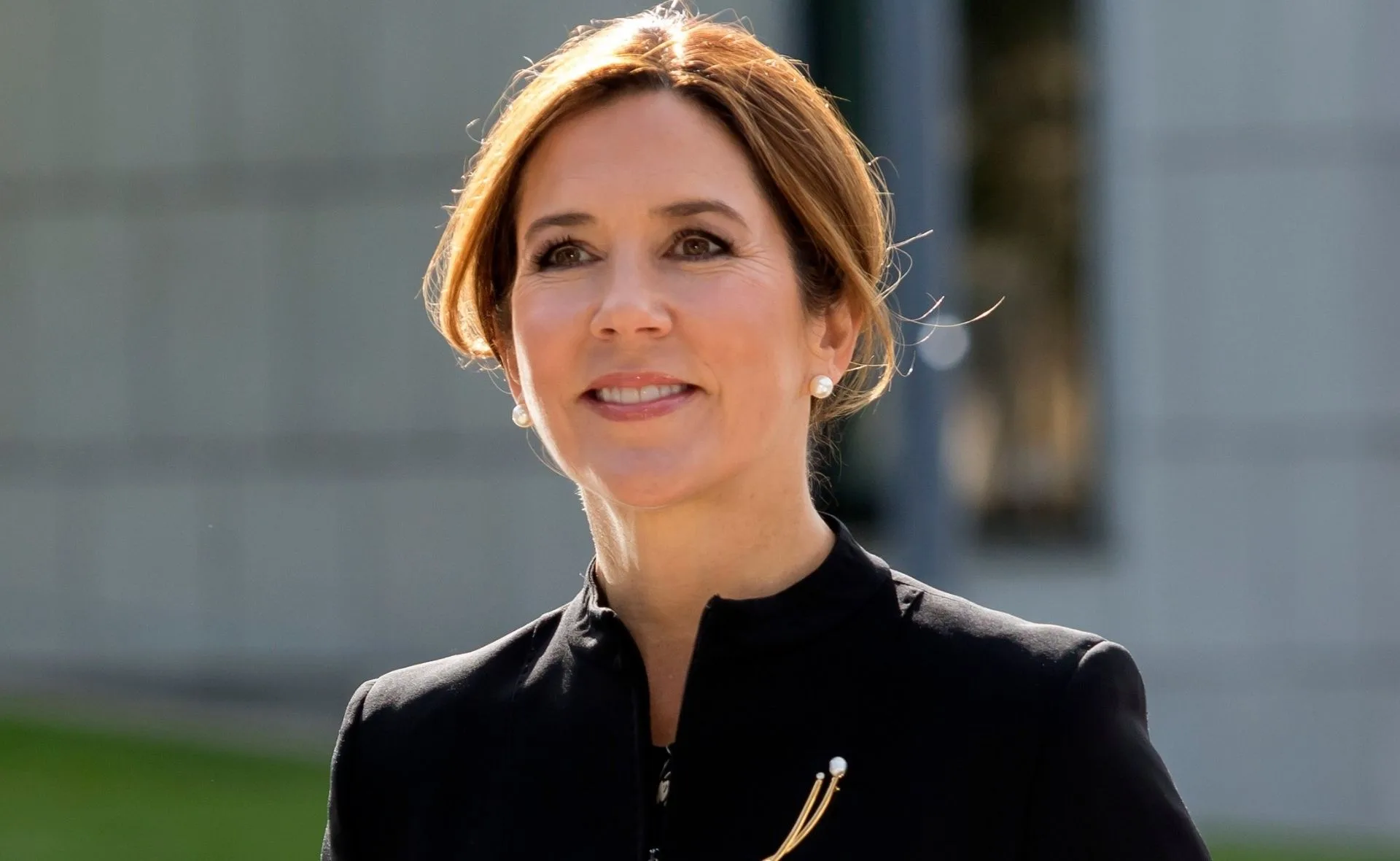A woman with earrings and a neat hairstyle smiles, wearing a black outfit with a gold brooch, against a blurred backdrop.