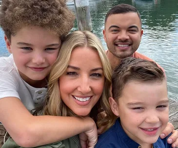 A family of four smiling in front of a waterfront, with two children hugging their parents, enjoying a sunny day outdoors.