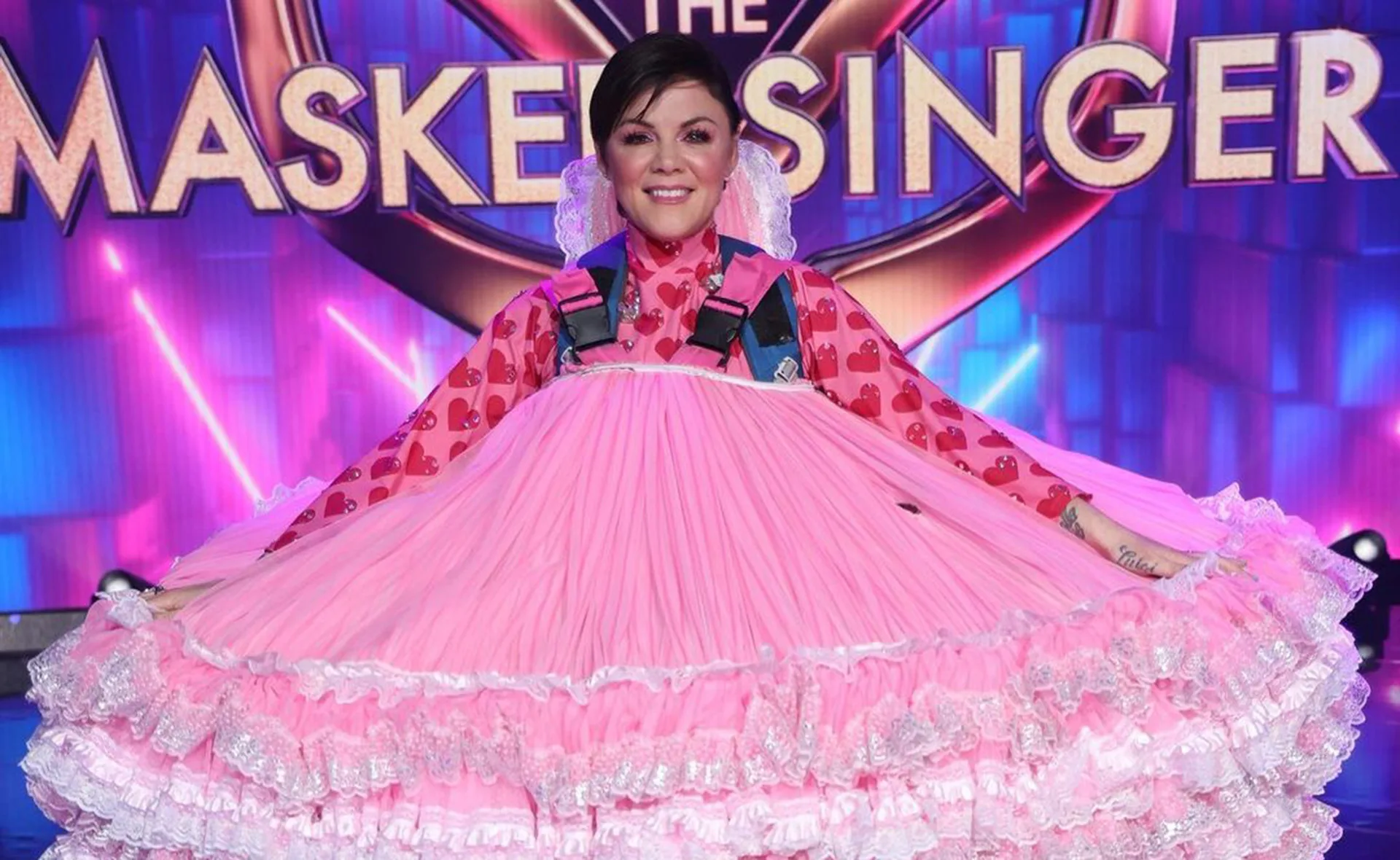 Person in pink heart-patterned dress, "The Masked Singer" logo in background.