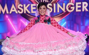 Person in pink heart-patterned dress, "The Masked Singer" logo in background.