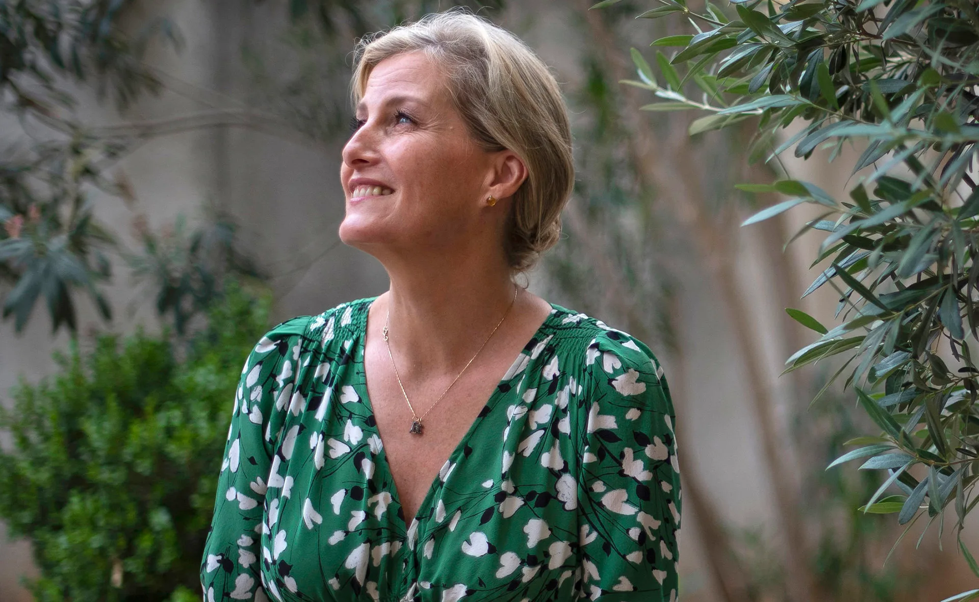A woman in a green floral dress smiles while looking up, surrounded by leafy plants.