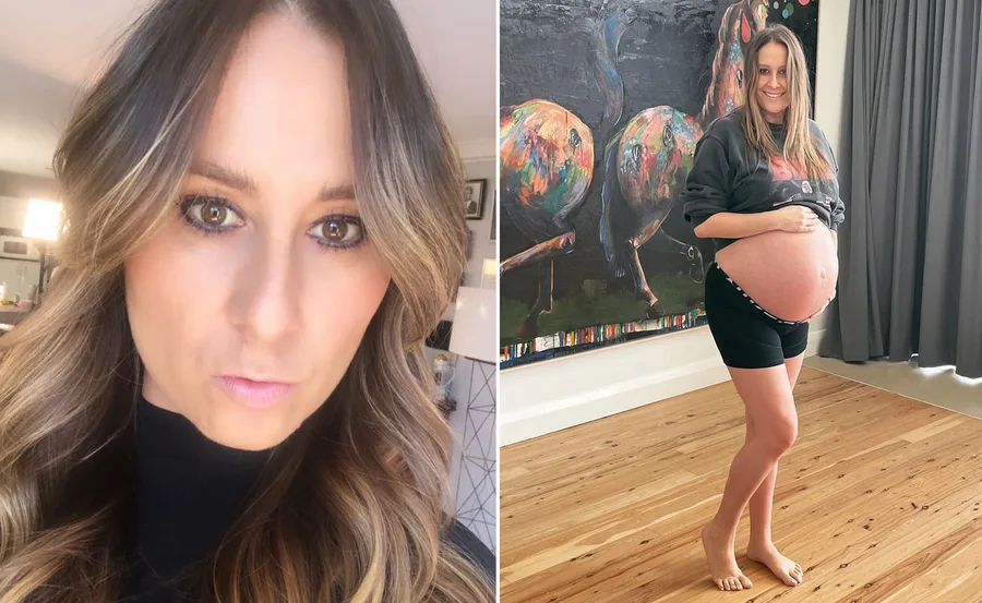 Pregnant woman in casual wear standing indoors, holding her belly, with a colorful abstract painting in the background.