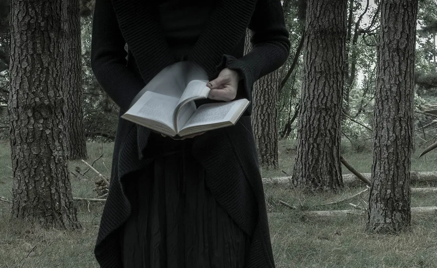 Person in dark clothing holding an open book in a forest setting.