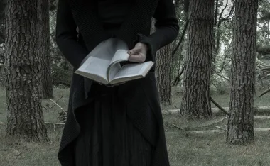Person in dark clothing holding an open book in a forest setting.