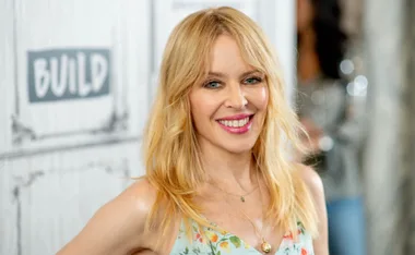 A person with long blonde hair smiles wearing a floral top, with a blurred background featuring a "BUILD" sign.