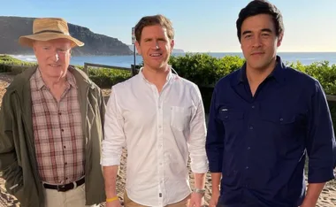 Three men standing on a beach with ocean and cliffs in the background, dressed casually in shirts.
