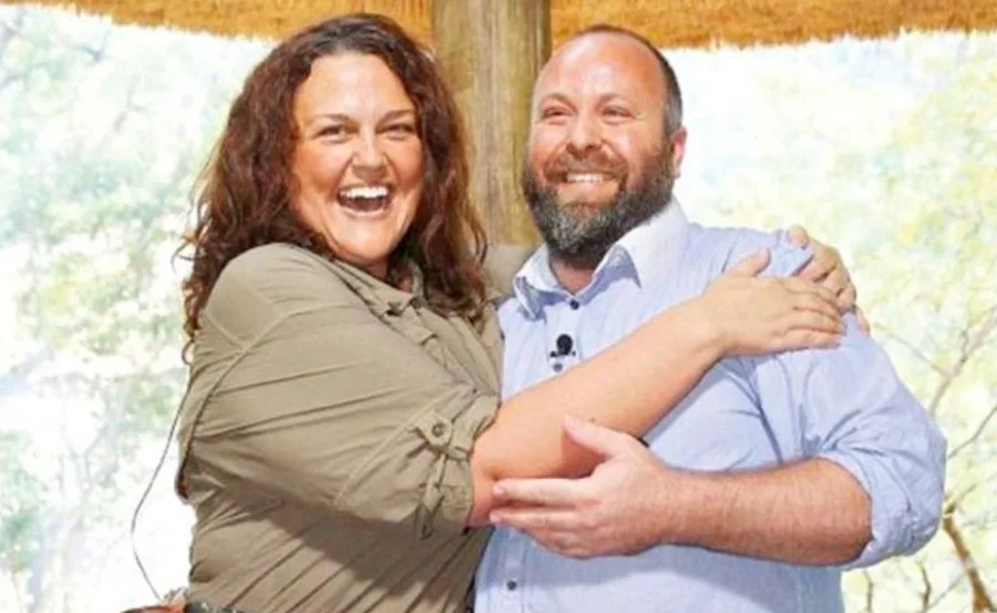 A smiling woman and man embrace outdoors under a thatched roof.