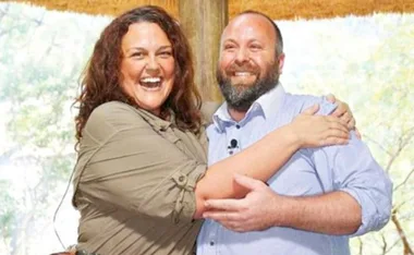 A smiling woman and man embrace outdoors under a thatched roof.