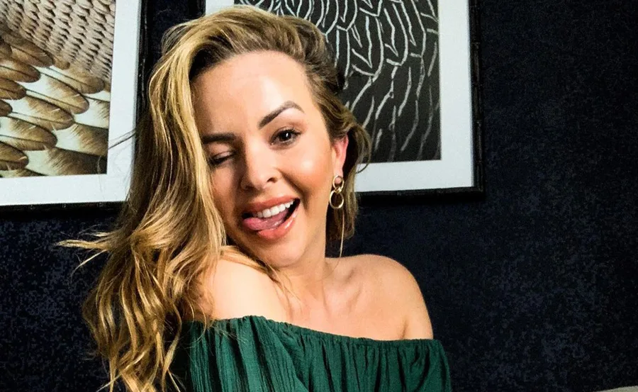 Person winking and smiling, wearing a green off-the-shoulder top, in front of abstract framed art.