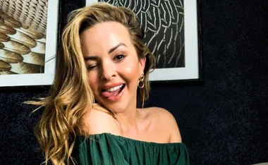 Person winking and smiling, wearing a green off-the-shoulder top, in front of abstract framed art.