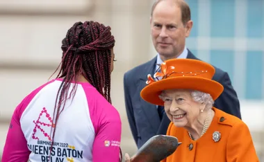 Queen in an orange outfit with a baton, smiling at a Baton Relay participant; a man in a suit stands nearby.