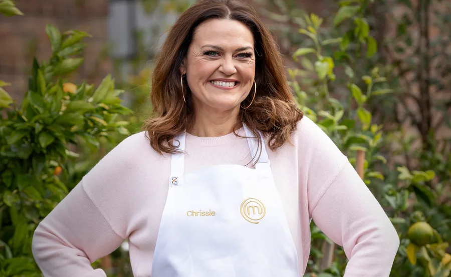 Chrissie Swan smiling in a "MasterChef" apron outdoors, surrounded by greenery.