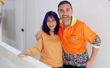 A woman with purple hair and a man in an orange shirt smiling and posing indoors.