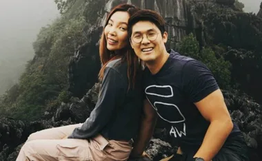 A couple smiling while sitting on rocky terrain in a misty, lush mountainous area.