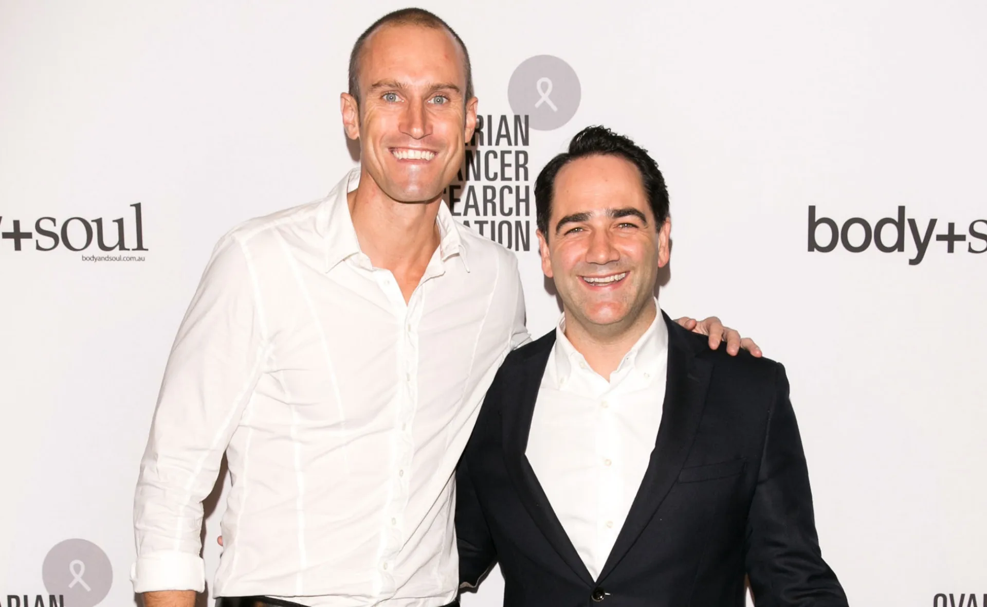 Two smiling men in white shirts at an event, standing in front of a white backdrop with logos.