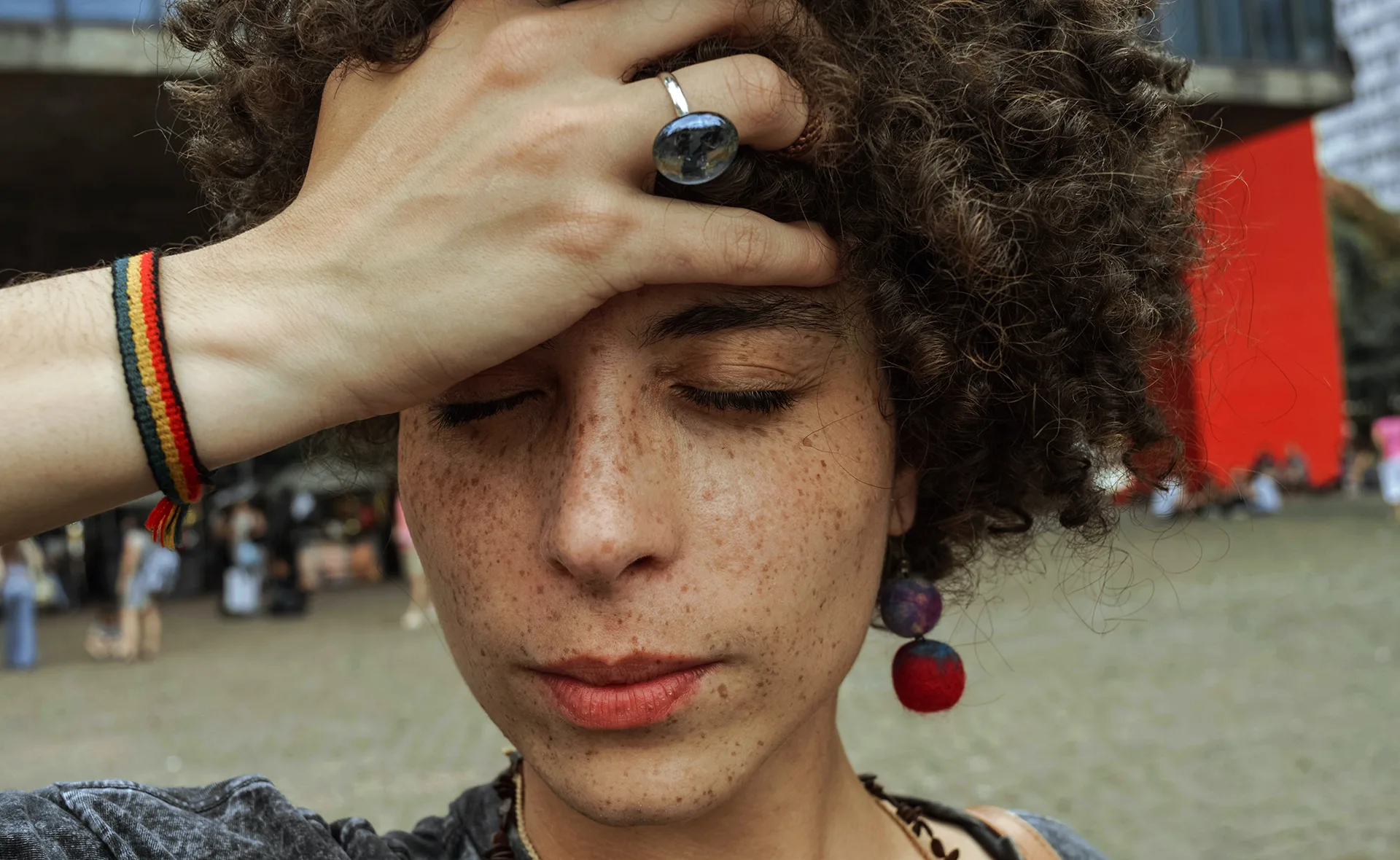 A person with curly hair and closed eyes, hand on forehead, wearing a ring and a multicolored bracelet.