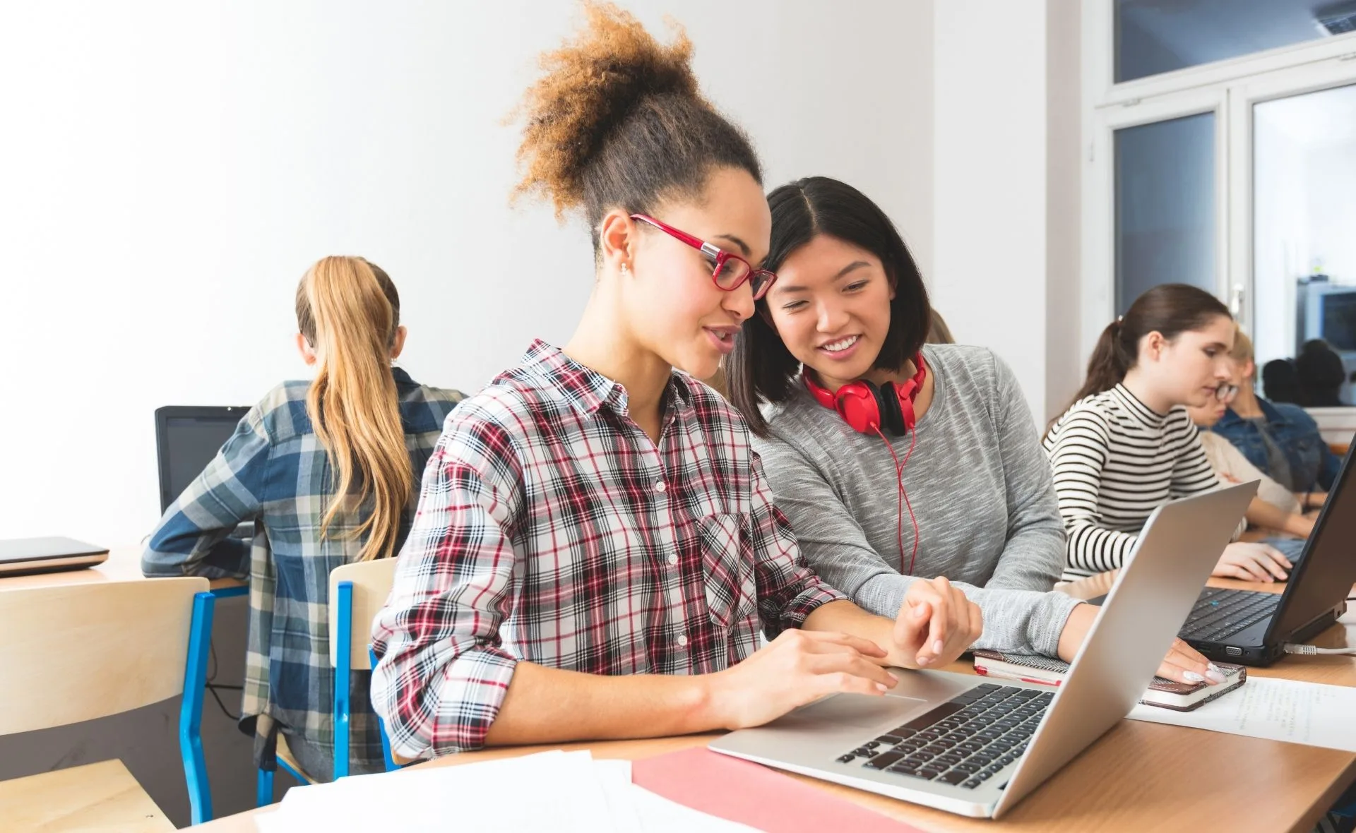 Two girls smiling and working on a laptop in a classroom setting, promoting tech education for girls.