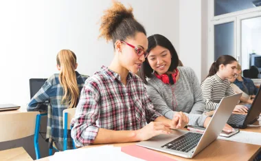 Two girls smiling and working on a laptop in a classroom setting, promoting tech education for girls.