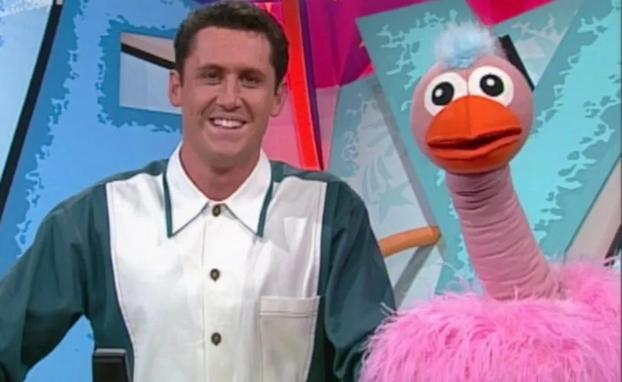 A man smiling beside a pink, fluffy puppet with a long neck and blue hair on a colorful set.