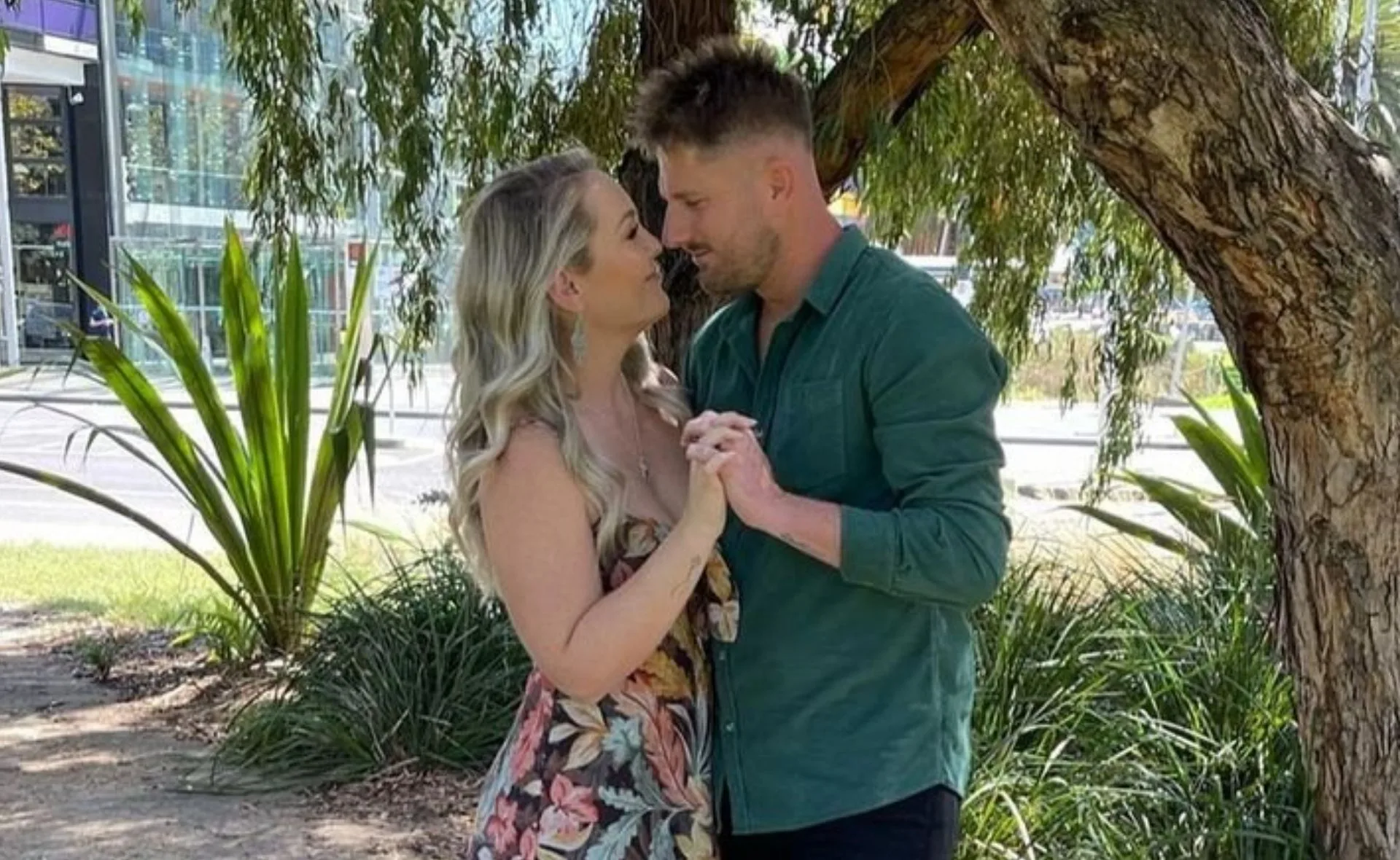 A couple holding hands and looking at each other lovingly under a tree in a park setting.