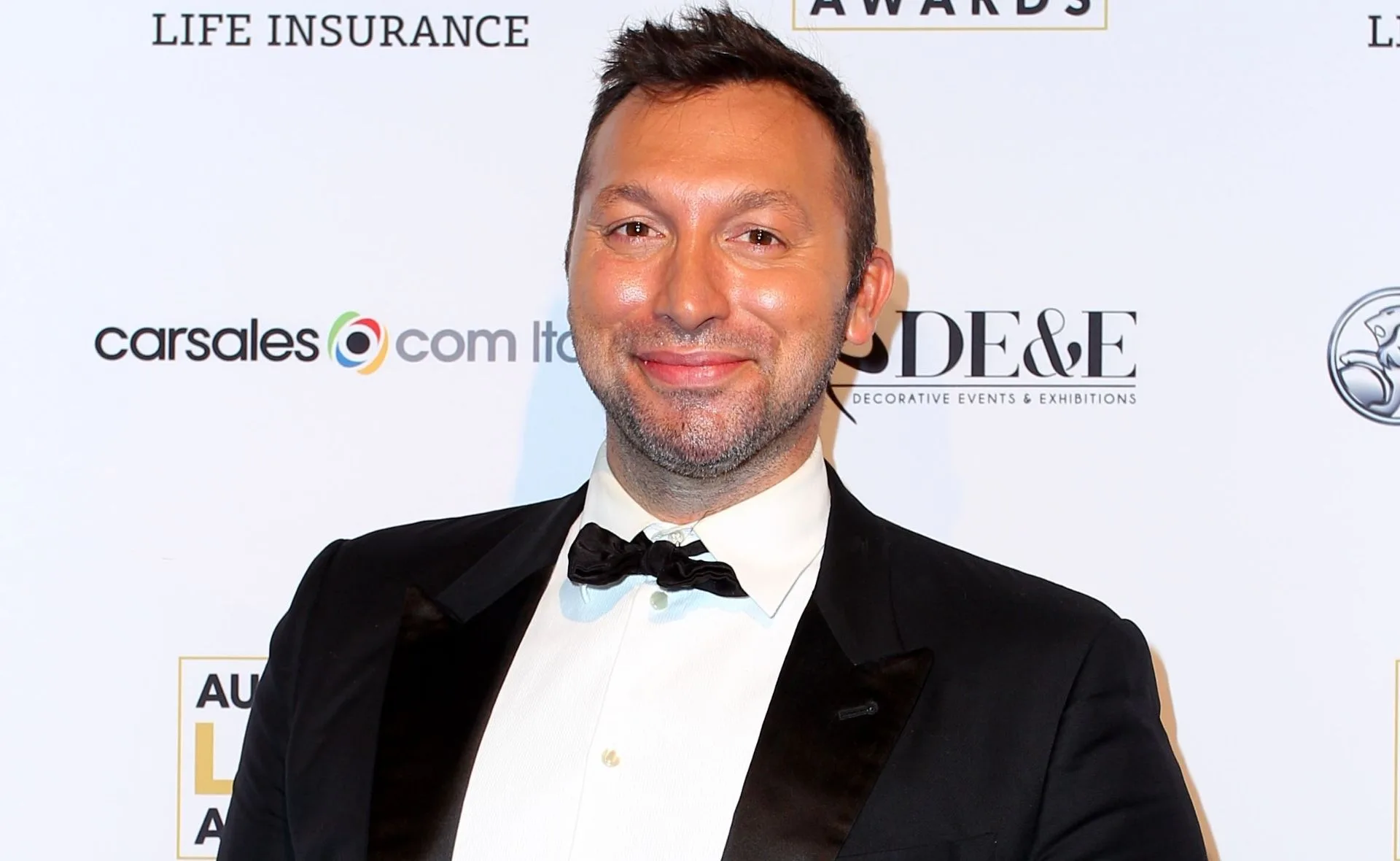 A person in a tuxedo smiling at an event with a branded backdrop.