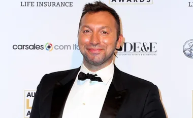 A person in a tuxedo smiling at an event with a branded backdrop.