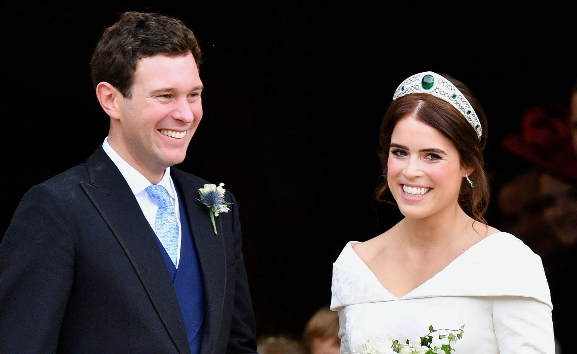 A couple smiling on their wedding day, the bride wearing a white gown and tiara, the groom in a suit with a boutonniere.