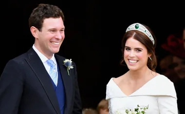 A couple smiling on their wedding day, the bride wearing a white gown and tiara, the groom in a suit with a boutonniere.