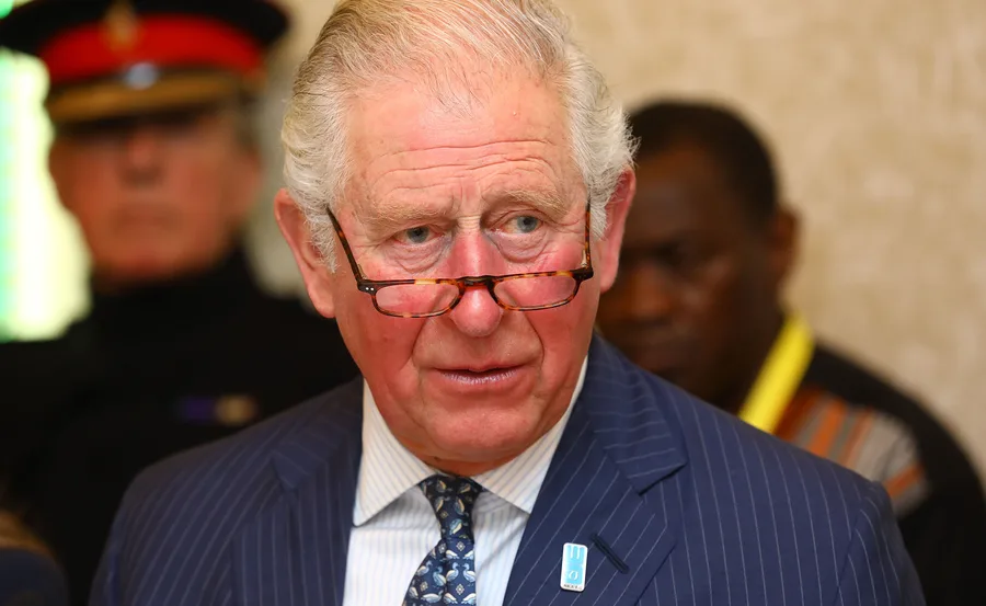 Prince Charles at an event, wearing glasses and a suit with a pin, a serious expression, people in the background.